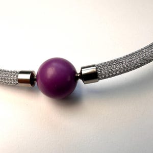 Op de afbeelding: Close-up van een zilveren mesh ketting met een levendige paarse bol in het midden. De ketting heeft zilveren metalen accenten. De achtergrond is effen wit.