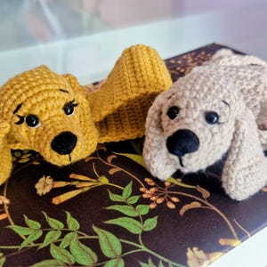 Puede incluir: Dos juguetes para perros hechos a mano con ganchillo. Uno es amarillo dorado, el otro beige claro. Ambos tienen ojos y nariz negros. Los perros están tumbados sobre un libro con un estampado floral.