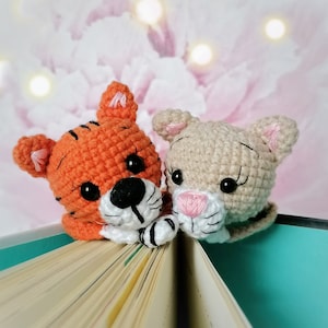 Könnte beinhalten: Zwei gehäkelte Tier-Lesezeichen, ein orange-weißes Tiger und eine beige Katze, liegen auf einem geöffneten Buch. Das Tiger-Lesezeichen hat schwarze Augen und eine schwarze Nase. Das Katzen-Lesezeichen hat schwarze Augen und eine rosa Nase.