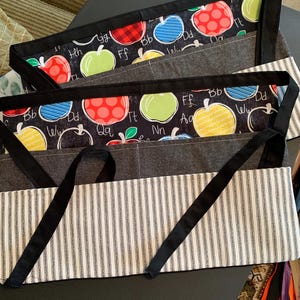 May include: Two aprons with black straps and pockets. The top section features a black background with colorful apple illustrations and letters. The bottom section has a white and black striped pattern.