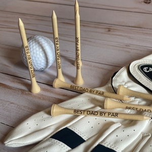 May include: A golf-themed gift set featuring wooden golf tees engraved with "BEST DAD BY PAR", a white golf ball, and a white golf glove with black accents. The items are arranged on a light wood surface.