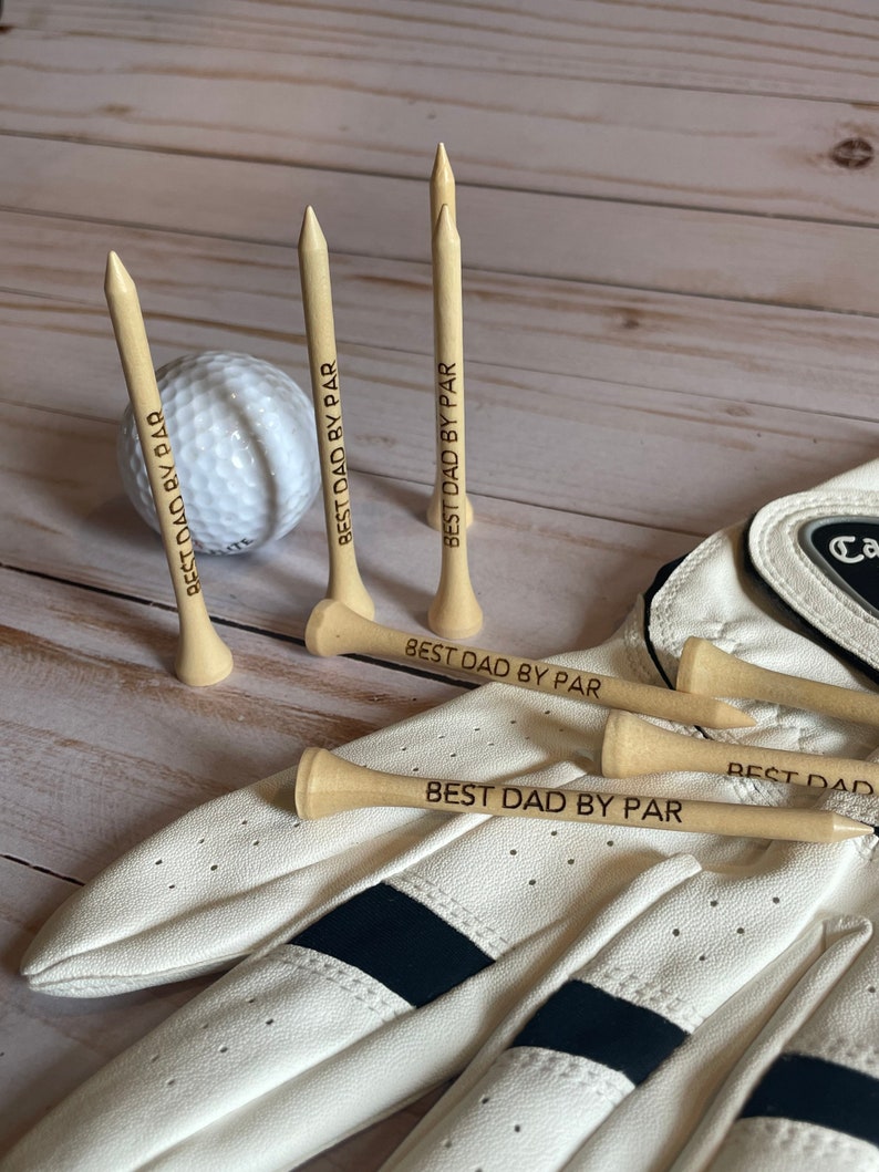 May include: A golf-themed gift set featuring wooden golf tees engraved with "BEST DAD BY PAR", a white golf ball, and a white golf glove with black accents. The items are arranged on a light wood surface.