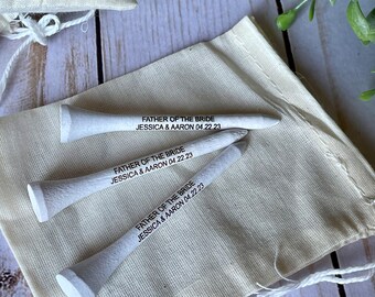 May include: Three white golf tees with the text "Father of the Bride Jessica & Aaron 04.22.23" printed on them. The tees are in a small white fabric bag.
