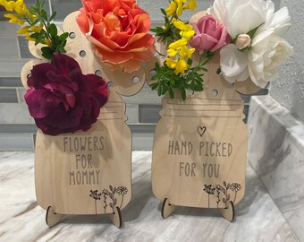 May include: Two wooden mason jar flower vases with stands. The vase on the left has the text "Flowers for Mommy" and the vase on the right has the text "Hand Picked for You".