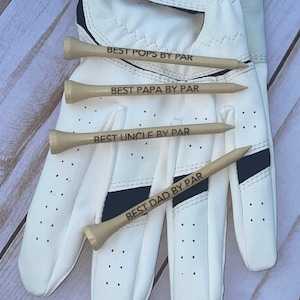 May include: Four wooden golf tees with the phrases "BEST POPS BY PAR", "BEST PAPA BY PAR", "BEST UNCLE BY PAR", and "BEST DAD BY PAR" engraved on them. The tees are arranged on a white golf glove with black accents.