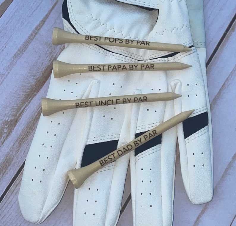 May include: Four wooden golf tees with the phrases "BEST POPS BY PAR", "BEST PAPA BY PAR", "BEST UNCLE BY PAR", and "BEST DAD BY PAR" engraved on them. The tees are arranged on a white golf glove with black accents.