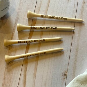 May include: Four wooden golf tees with the text "BEST DAD BY PAR" engraved on them. A golf ball and a golf glove are also visible. The tees are light brown and are arranged on a light wood surface.