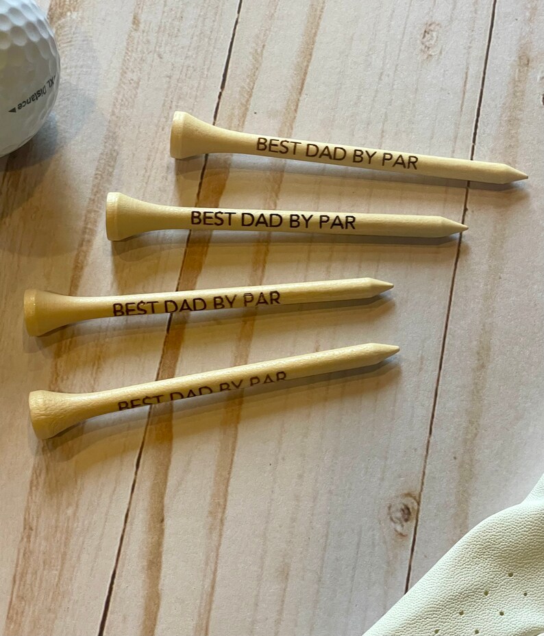 May include: Four wooden golf tees with the text "BEST DAD BY PAR" engraved on them. A golf ball and a golf glove are also visible. The tees are light brown and are arranged on a light wood surface.