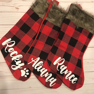 May include: Three red and black buffalo plaid Christmas stockings with faux fur trim. The stockings are personalized with the names "Rocky", "Aliana", and "Rance".