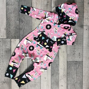 K&ouml;nnte beinhalten: Rosa-schwarzer Baby-Strampler mit einem Retro-Design aus den 80er Jahren mit Rollschuhen, Kassetten und Schallplatten. Der Strampler hat eine Kapuze.