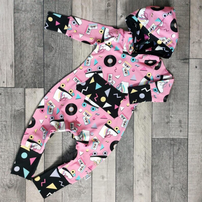 K&ouml;nnte beinhalten: Rosa-schwarzer Baby-Strampler mit einem Retro-Design aus den 80er Jahren mit Rollschuhen, Kassetten und Schallplatten. Der Strampler hat eine Kapuze.