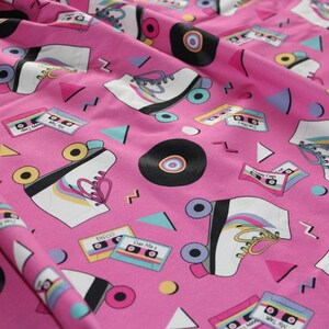 K&ouml;nnte beinhalten: Rosafarbener Stoff mit einem sich wiederholenden Muster aus Rollschuhen, Kassetten und Schallplatten. Die Rollschuhe sind wei&szlig; mit regenbogenfarbenen Rollen und Schn&uuml;rsenkeln. Die Kassetten haben verschiedene Titel, darunter "Star Mix 2", "Happy Camp" und "Mr. Jones's Album".