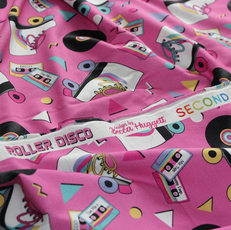 K&ouml;nnte beinhalten: Rosafarbener Stoff mit einem sich wiederholenden Muster aus Rollschuhen, Kassetten und Schallplatten. Der Stoff hat einen wei&szlig;en Rand mit dem Text "ROLLER DISCO" und "design by Becca Higgett SECOND".