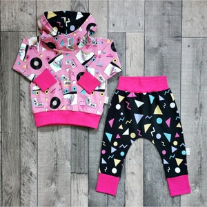 K&ouml;nnte beinhalten: Ein rosa-schwarzes Baby-Outfit mit einem Kapuzenpullover und einer passenden Hose. Der Pullover hat ein Retro-Design aus den 80ern mit Rollschuhen, Kassetten und Schallplatten. Die Hose hat ein geometrisches Muster mit Dreiecken, Kreisen und Zickzacklinien.