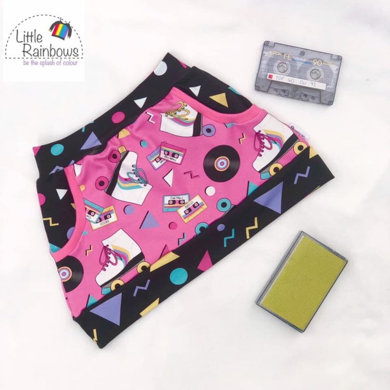 K&ouml;nnte beinhalten: Pinkfarbener Stoff mit einem geometrischen Muster in Schwarz, Wei&szlig; und Blau. Der Stoff zeigt ein sich wiederholendes Muster aus Rollschuhen, Kassetten und Schallplatten. Der Text "Little Rainbows" ist in der oberen linken Ecke des Bildes sichtbar.