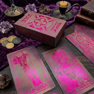 Könnte beinhalten: Eine Tarot-Kartenschachtel mit der Aufschrift MAGNIFIQUE TAROT DECK in metallisch-pinker Folie auf dunkelbraunem Grund. Zwei Tarotkarten liegen offen da und zeigen den Eremiten und den Wagen in passender pinker Folie auf braunem Karton.