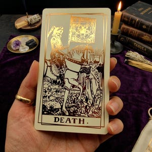 May include: A rectangular tarot card with a cream background and metallic gold foil illustration. The design features a skeleton in armor riding a horse, holding a flag with a rose emblem. The word DEATH is printed in gold at the bottom.