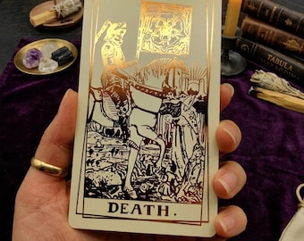 Può includere: Una carta dei tarocchi rettangolare con sfondo color crema e illustrazione in lamina d'oro metallizzata. Il disegno raffigura uno scheletro in armatura a cavallo che regge una bandiera con un emblema a forma di rosa. La parola DEATH è stampata in oro.