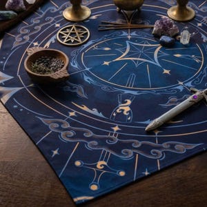 Puede incluir: Un mantel de altar de tela azul oscuro con un diseño circular en dorado y azul claro que presenta motivos celestiales, nubes e imágenes de espadas. El mantel está dispuesto con un cuenco de madera con hierbas, un pentáculo de latón, cristales y una daga.