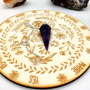 May include: A wooden pendulum board with a floral design and the words "Yes", "Maybe", "Rephrase", and "No". A purple amethyst pendulum is hanging over the board.
