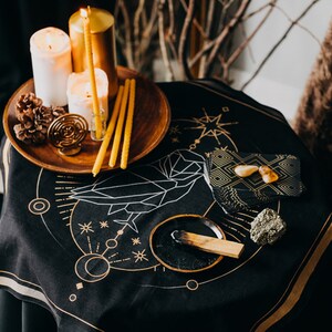 May include: A black altar cloth with a gold geometric design, featuring candles, a pine cone, a wooden bowl with a burning stick of incense, and a small stack of black slate tiles with gold accents.