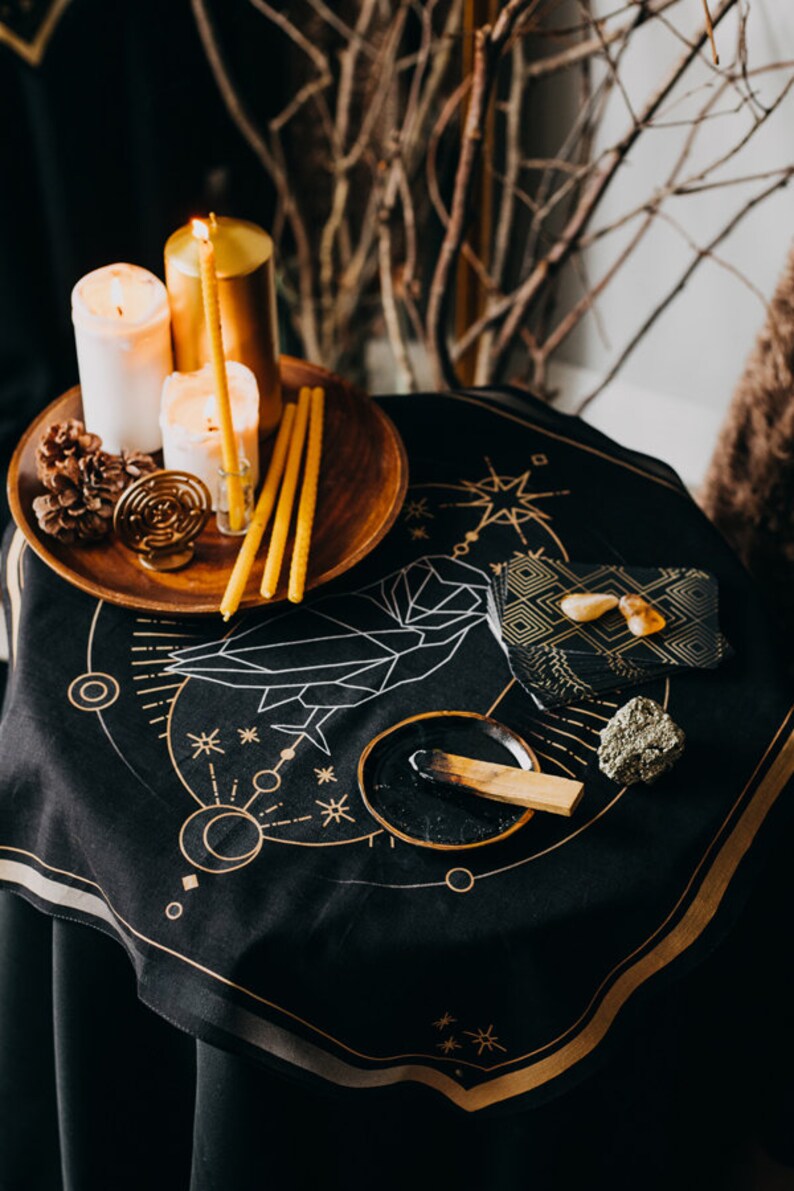 May include: A black altar cloth with a gold geometric design, featuring candles, a pine cone, a wooden bowl with a burning stick of incense, and a small stack of black slate tiles with gold accents.