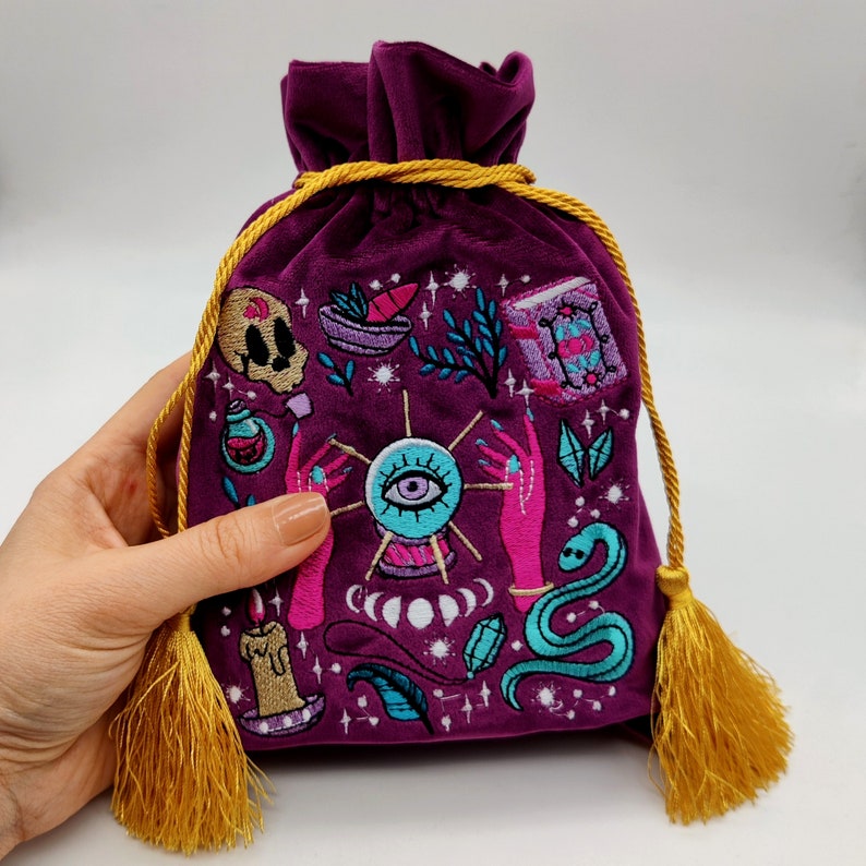 May include: Purple velvet pouch with a gold drawstring and tassel. The pouch is embroidered with a variety of colorful images, including an eye, hands, a snake, a skull, and a book.