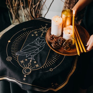 May include: A black circular tapestry with a gold geometric owl design and a gold border. A wooden tray with three candles and pine cones sits on top of the tapestry.
