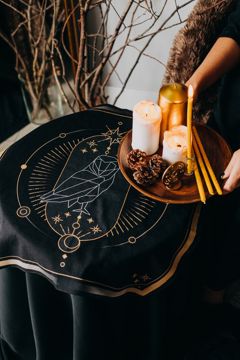 May include: A black circular tapestry with a gold geometric owl design and a gold border. A wooden tray with three candles and pine cones sits on top of the tapestry.