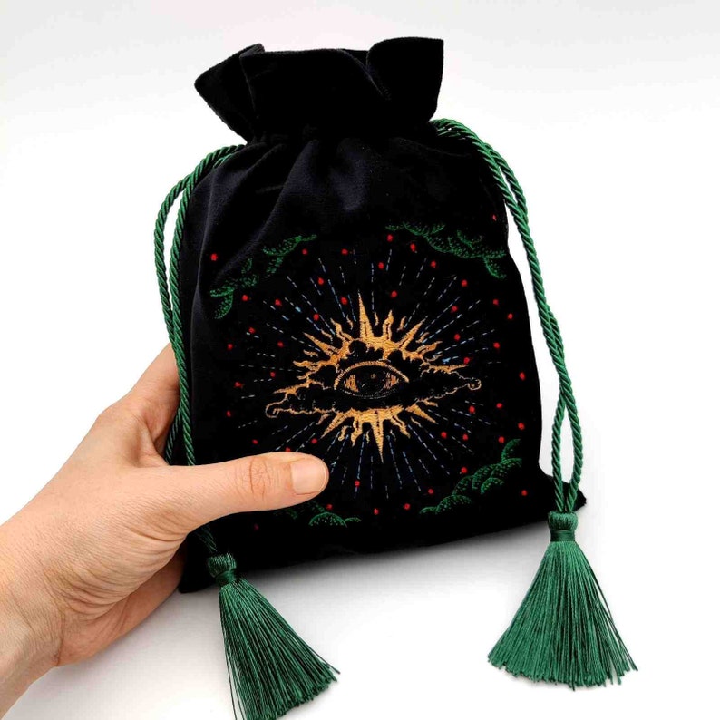 May include: Black velvet drawstring bag with an embroidered gold sunburst design featuring an eye. The bag has green drawstrings and tassels. The design includes red and green accents.
