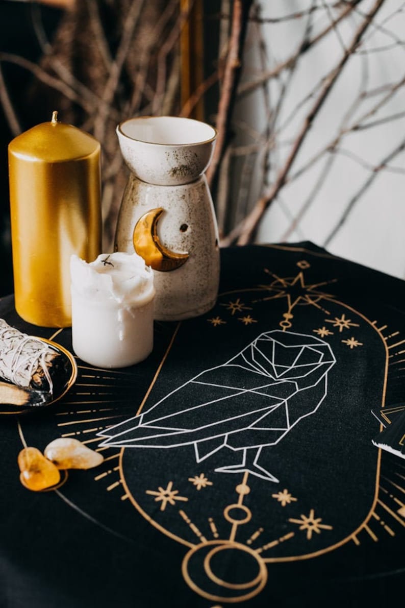 May include: A black altar cloth with gold geometric designs, including an owl, stars, and a crescent moon. There are three candles, two white and one gold, and a white ceramic oil burner with a gold crescent moon design.