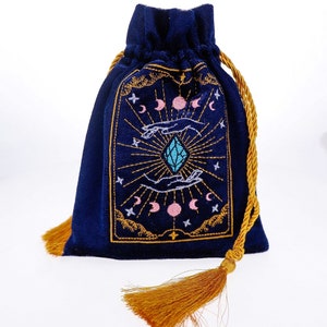 May include: A dark blue velvet pouch with a gold drawstring and tassel. The pouch features an embroidered design of a crystal, hands, and moons.
