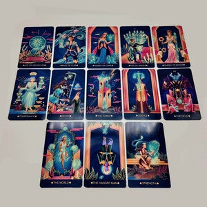 May include: A set of 14 tarot cards with colorful illustrations and gold accents. The cards feature characters and symbols from the Major Arcana, including The World, The Hanged Man, Strength, Temperance, Death, The Tower, Justice, The Devil, Queen of Wands, Ten of Wands, Queen of Cups, Page of Cups, and Ace of Pentacles.