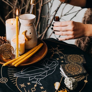May include: A black table cloth with gold geometric designs and a white owl illustration. There are candles, a diffuser, and a bottle of essential oil on the table.