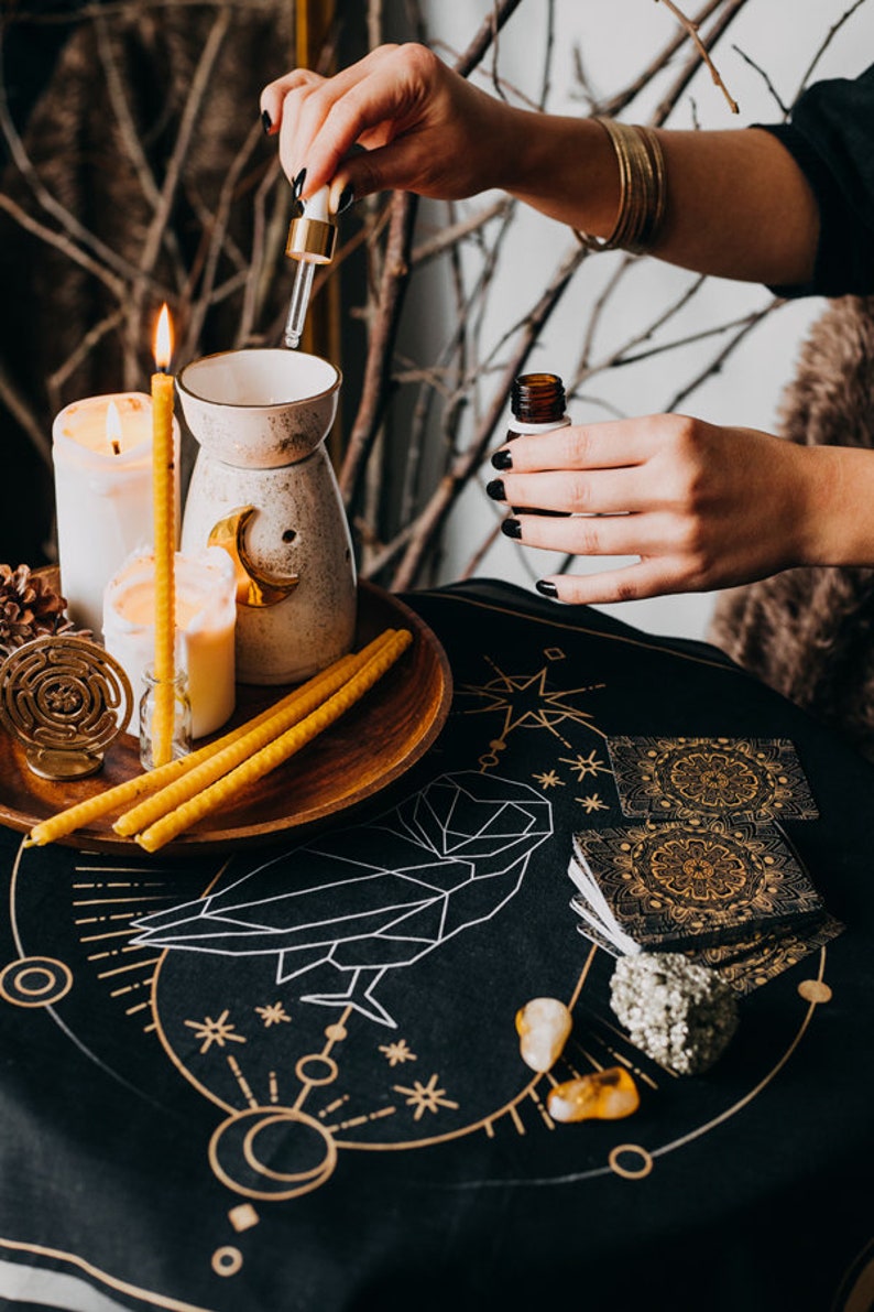 May include: A black table cloth with gold geometric designs and a white owl illustration. There are candles, a diffuser, and a bottle of essential oil on the table.