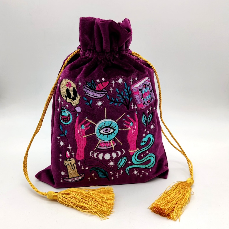 May include: Purple velvet drawstring bag with a gold tassel. The bag is embroidered with a colorful design featuring an eye, a snake, a candle, and other mystical symbols.