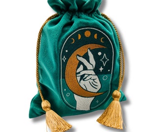 May include: Teal velvet drawstring bag with gold tassels. The bag features an embroidered design of a hand holding a crescent moon with stars and the word "DOC"