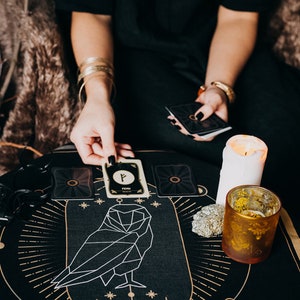May include: A black tarot cloth with a geometric owl design and gold accents. A white candle and a gold glass are on the cloth. A person is holding a tarot card with the rune "Fehu" on it.