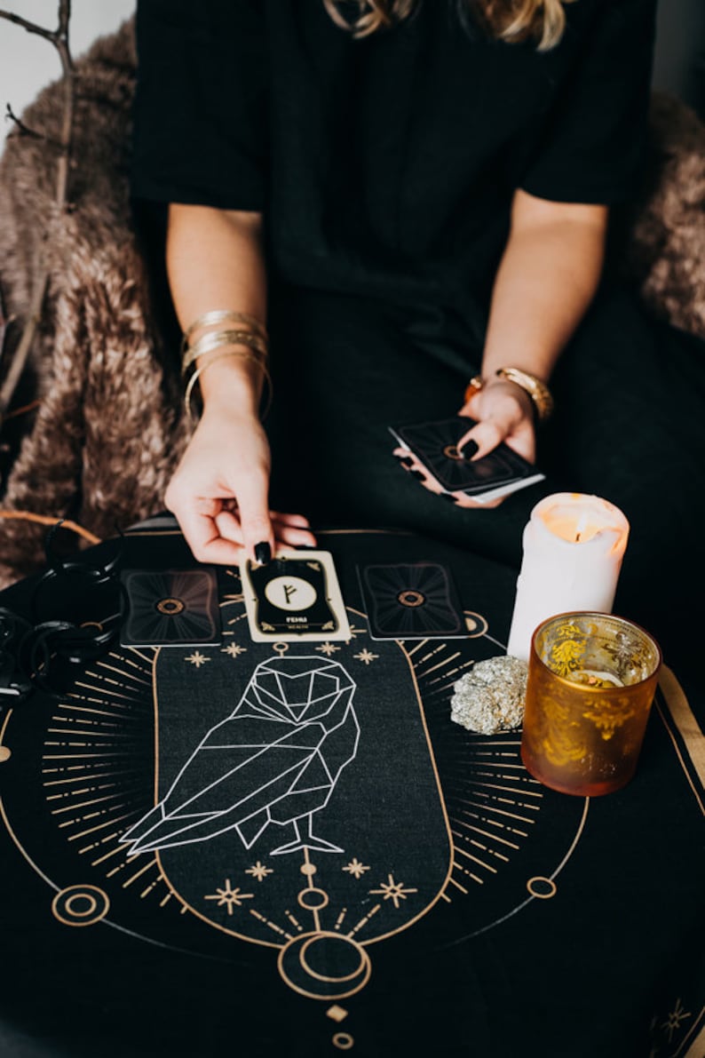 May include: A black tarot cloth with a geometric owl design and gold accents. A white candle and a gold glass are on the cloth. A person is holding a tarot card with the rune "Fehu" on it.