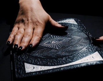 May include: A black leather-bound journal with an embossed silver eye design on the cover. The journal is closed and lying flat on a dark surface.