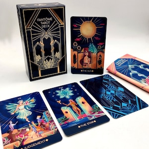 May include: A tarot deck box with gold accents and the text "FANT&Ocirc;ME TAROT DECK". The box is closed and sitting on a white surface. There are several tarot cards spread out around the box, including the "The Sun", "The Star", and "Judgement" cards.