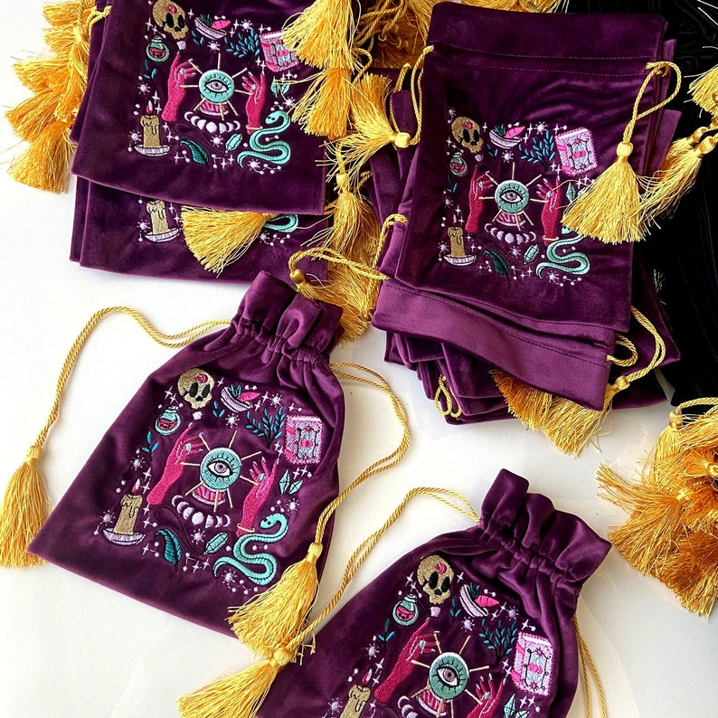 May include: A stack of purple velvet pouches with gold tassels. Each pouch features an embroidered design of an eye, hands, a snake, and other mystical symbols.