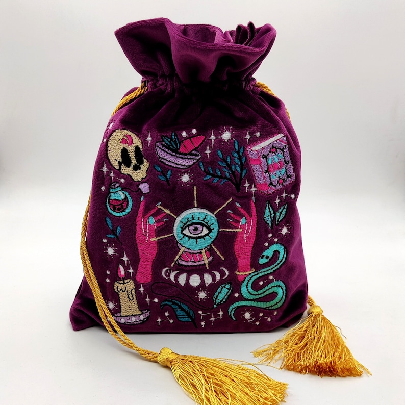 May include: A purple velvet pouch with a gold drawstring and tassel. The pouch is embroidered with a variety of images, including an eye, a skull, a book, and a snake.