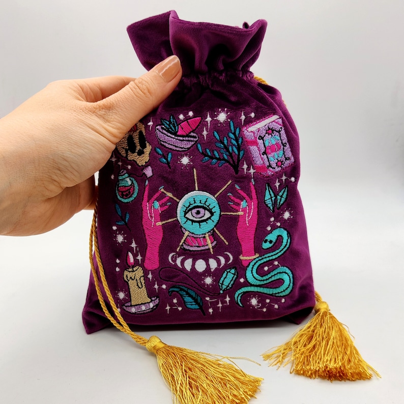 May include: A purple velvet pouch with a drawstring closure and gold tassels. The pouch is embroidered with a variety of colorful images, including an eye, a snake, a candle, and a hand holding a crystal ball. The pouch is perfect for storing small items like jewelry, tarot cards, or crystals.
