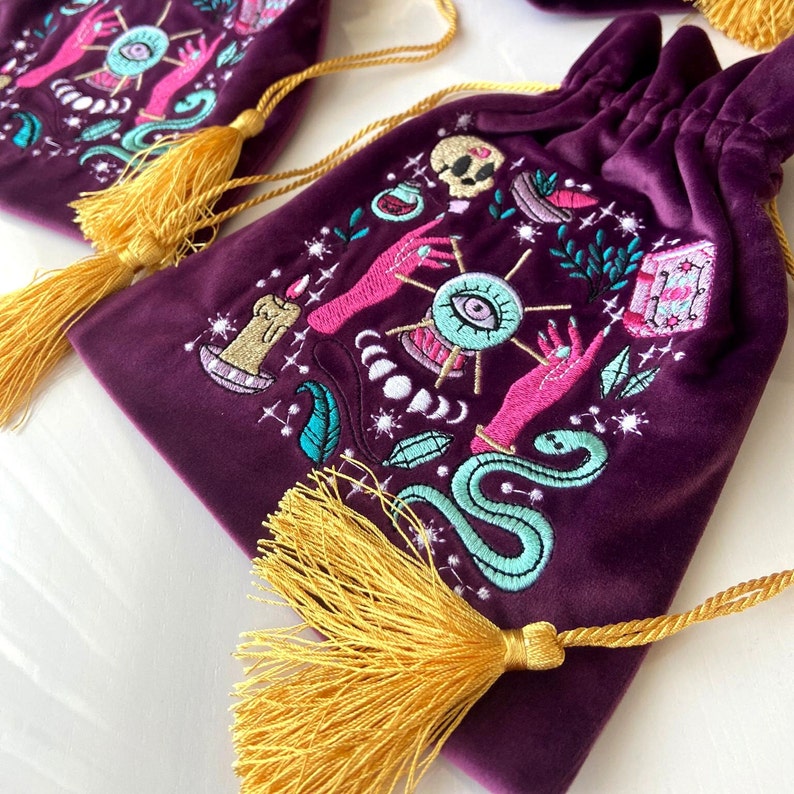 May include: Purple velvet drawstring bag with gold tassels and embroidered details of a snake, an eye, a hand, and a candle. The bag has a mystical and magical theme.