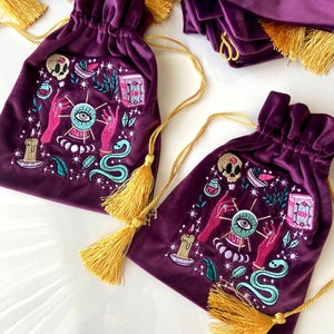 May include: Two purple velvet drawstring bags with gold tassels. Each bag features an embroidered design of a mystical eye, hands, a snake, a candle, and other magical symbols.