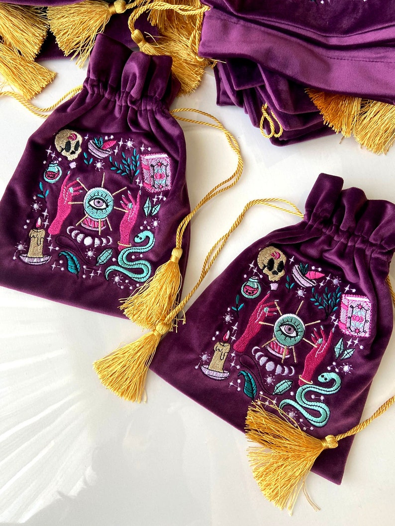 May include: Two purple velvet drawstring bags with gold tassels. Each bag features an embroidered design of a mystical eye, hands, a snake, a candle, and other magical symbols.