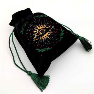 May include: Black velvet drawstring bag with a gold and white embroidered eye design, surrounded by a sunburst pattern. The bag has green drawstrings and tassels, perfect for storing small items or as a unique gift.