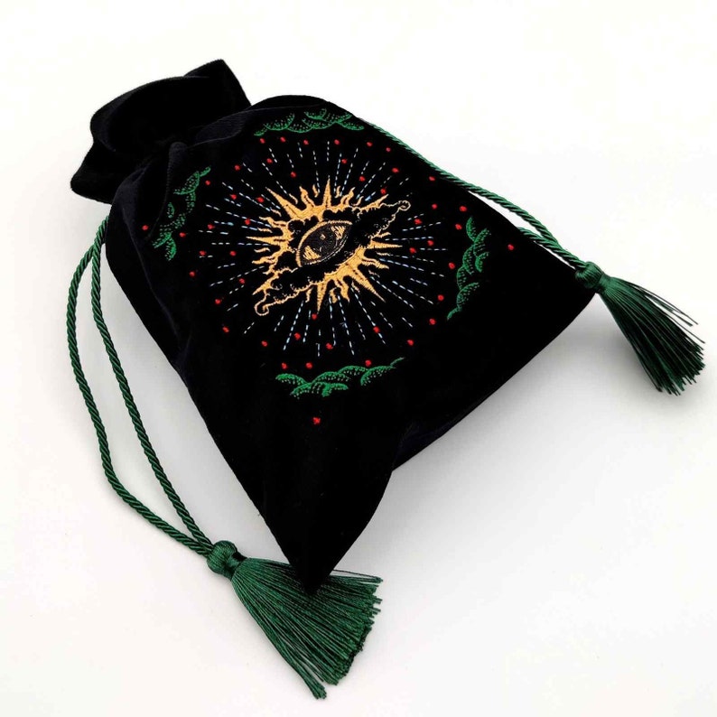 May include: Black velvet drawstring bag with a gold and white embroidered eye design, surrounded by a sunburst pattern. The bag has green drawstrings and tassels, perfect for storing small items or as a unique gift.