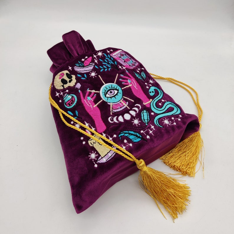 May include: A purple velvet drawstring bag with a gold tassel. The bag is embroidered with a colorful design of an eye, hands, a snake, and other mystical symbols.